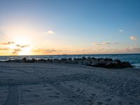 the sun sets on an empty beach with rocks and sand in front of it,