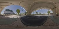 a 360 - spherical photo taken under a building with arches, the view is from a street level in which two people are sitting on a bench and walking