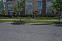 a paved city street next to two apartment buildings with tall windows and a green lawn