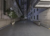 an empty sidewalk is in between two tall buildings with lights on the windows and a small bench at the end of it