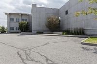 an empty parking lot with grass growing out of it's corner and a tree in front of a large, gray concrete building