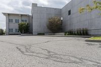 an empty parking lot with grass growing out of it's corner and a tree in front of a large, gray concrete building