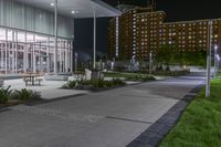 some benches and lights near the building at night's edge of park and sidewalk