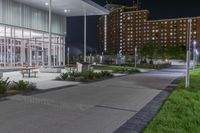 some benches and lights near the building at night's edge of park and sidewalk