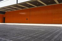 an orange wall next to a white building on brick paving on a sidewalk in front of a walkway