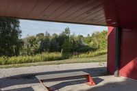 a wooden bench sits beneath a roof on a cement lot in the shade, next to some trees and grass