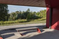 a wooden bench sits beneath a roof on a cement lot in the shade, next to some trees and grass