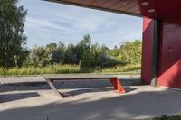 a wooden bench sits beneath a roof on a cement lot in the shade, next to some trees and grass