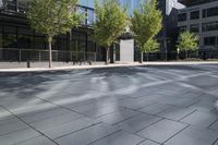 two people are walking on an empty city sidewalk by some buildings, and several trees, and benches