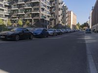 cars lined up in a row on the side of a street, along with apartment blocks lining either side of the road