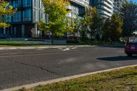 a red car drives past two large modern buildings in the background, with autumn trees