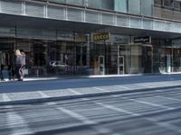 a person walking down a sidewalk in front of a building with a storefront and car in the background