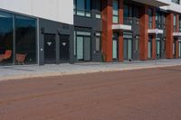 there are two wooden chairs near the street corner of this city street building, and a red brick sidewalk to the right