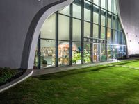 a building with large windows on top of a lush green lawn outside at night time