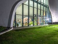 a building with large windows on top of a lush green lawn outside at night time