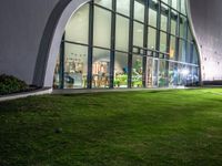 a building with large windows on top of a lush green lawn outside at night time