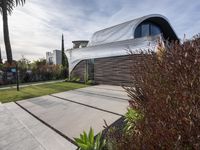 the modern house features curved roof and stone pavers in its backyard area with palm trees on one side and greenery on the other