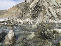 the rocks and pebbles at the edge of the water are surrounded by an eroded cliff