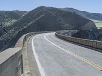 a motorcycle is driving along the curved road over a bridge in a valley on a sunny day