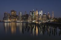 the city skyline at night with reflection in water surface and wooden pillars on shore,