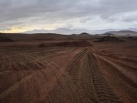 a dirt road through a desert area with mountains in the distance in a cloudy sky