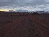 a dirt road through a desert area with mountains in the distance in a cloudy sky