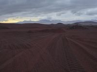 a dirt road through a desert area with mountains in the distance in a cloudy sky