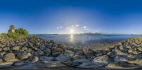 some rocks and the sun is setting on a beach with a water view of city skyline