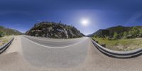 a fish eye lens panoramic view of a road surrounded by mountains, trees, and grass