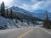 a snow covered road and mountain with evergreen trees on both sides of it, and an empty field and road to the left