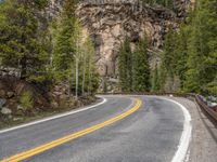 the curved road curves in front of a mountain area with pine trees behind it,