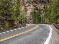 the curved road curves in front of a mountain area with pine trees behind it,