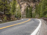 the curved road curves in front of a mountain area with pine trees behind it,