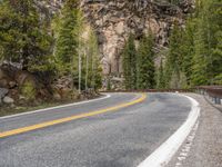 the curved road curves in front of a mountain area with pine trees behind it,