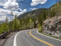 the curved road curves in front of a mountain area with pine trees behind it,