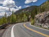 the curved road curves in front of a mountain area with pine trees behind it,