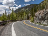 the curved road curves in front of a mountain area with pine trees behind it,