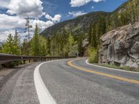 the curved road curves in front of a mountain area with pine trees behind it,