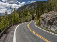 the curved road curves in front of a mountain area with pine trees behind it,