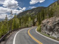 the curved road curves in front of a mountain area with pine trees behind it,