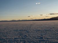 a field filled with white snow next to mountains at sunset in a blue sky with a few white clouds