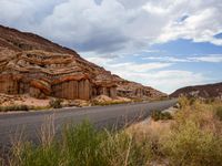 the side of an empty road is surrounded by vegetation and a rocky cliff behind it