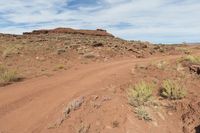 a dirt road in a dry landscape with brush and rocks in the foreground and a rock formation to the right