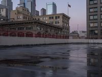 an empty parking lot with some buildings in the background and water on it and a flag pole overhanging the sidewalk