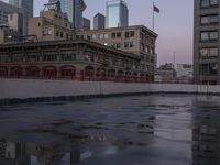 an empty parking lot with some buildings in the background and water on it and a flag pole overhanging the sidewalk