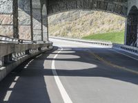 a motorcycle is riding down a road under a bridge a large mountain has grass and shrubs