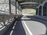 a motorcycle is riding down a road under a bridge a large mountain has grass and shrubs