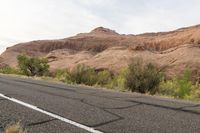 the road runs through the desert and there are hills in the distance, there is a large stone - covered mountain behind