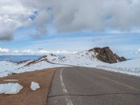 an empty road with snow and mountains in the back ground under cloudy skies on a sunny day