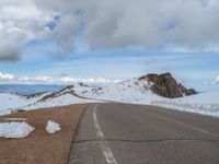 an empty road with snow and mountains in the back ground under cloudy skies on a sunny day